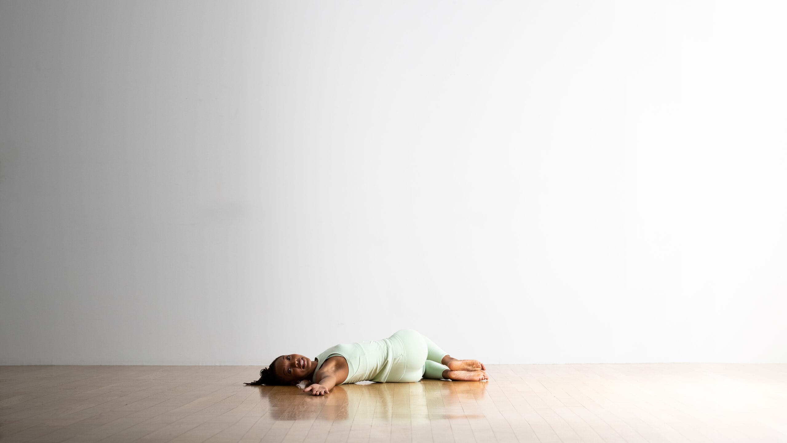 Young woman wearing light green top and tights is lying down to practice Supta Matsyendrasana (Supine Spinal Twist)