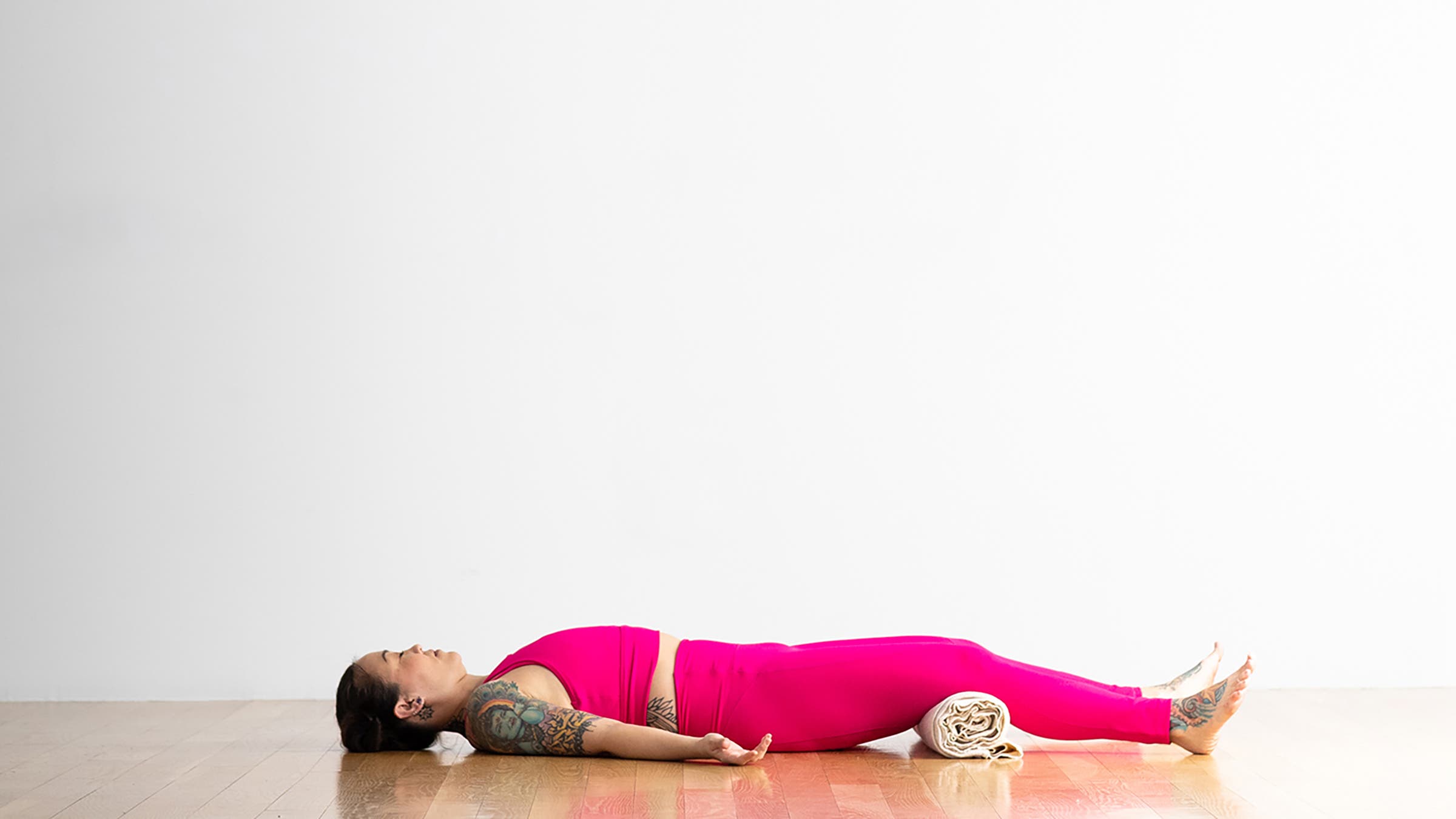 A person demonstrates a variation of Savasana (Corpse Pose) in yoga, with a rolled blanket under the knees