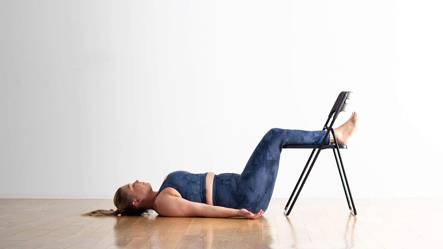 A person demonstrates a variation of Savasana (Corpse Pose) in yoga, with their feet up on a chair