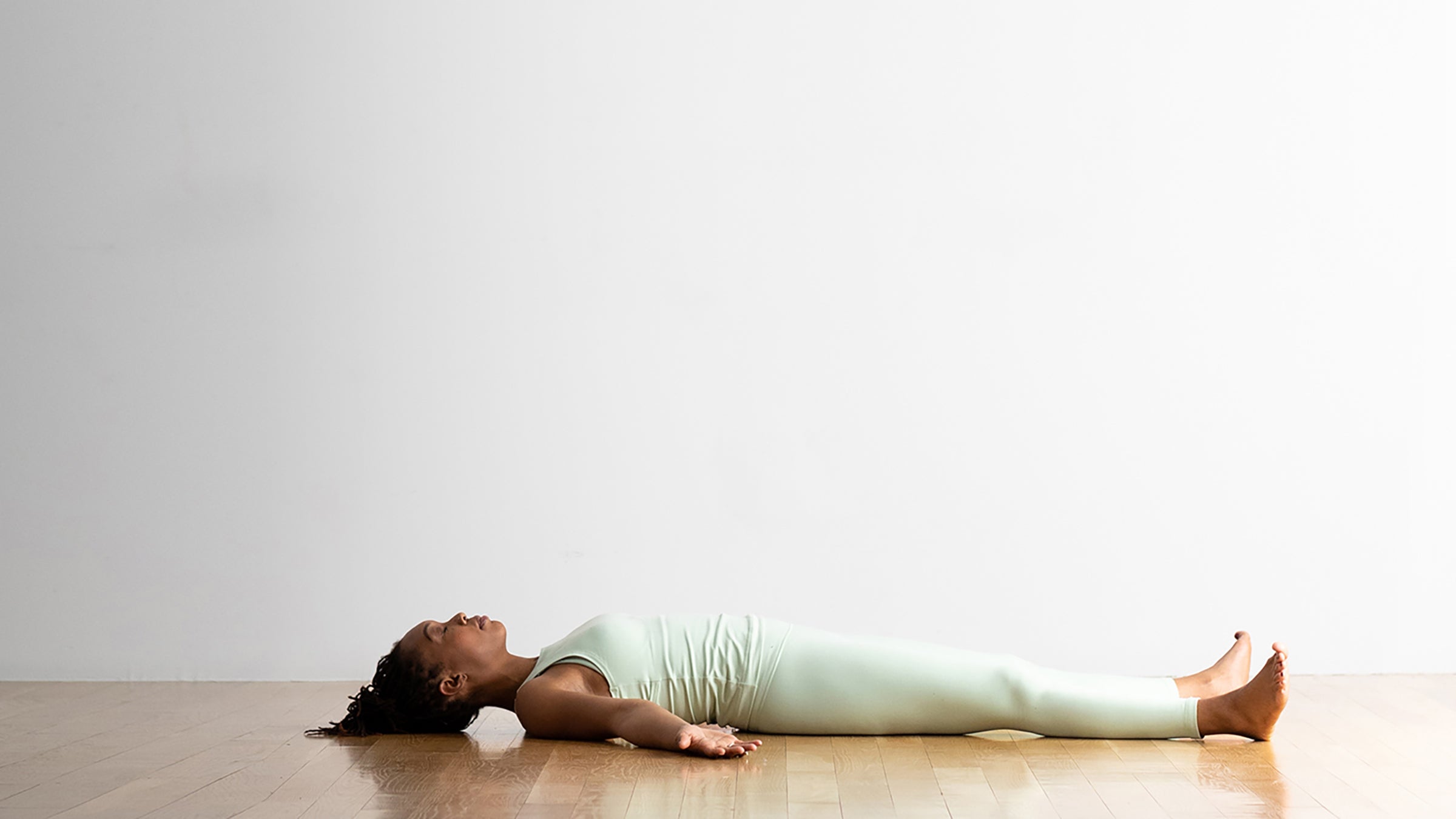 A Black woman in sea-green clothes person demonstrates Savasana (Corpse Pose) in yoga