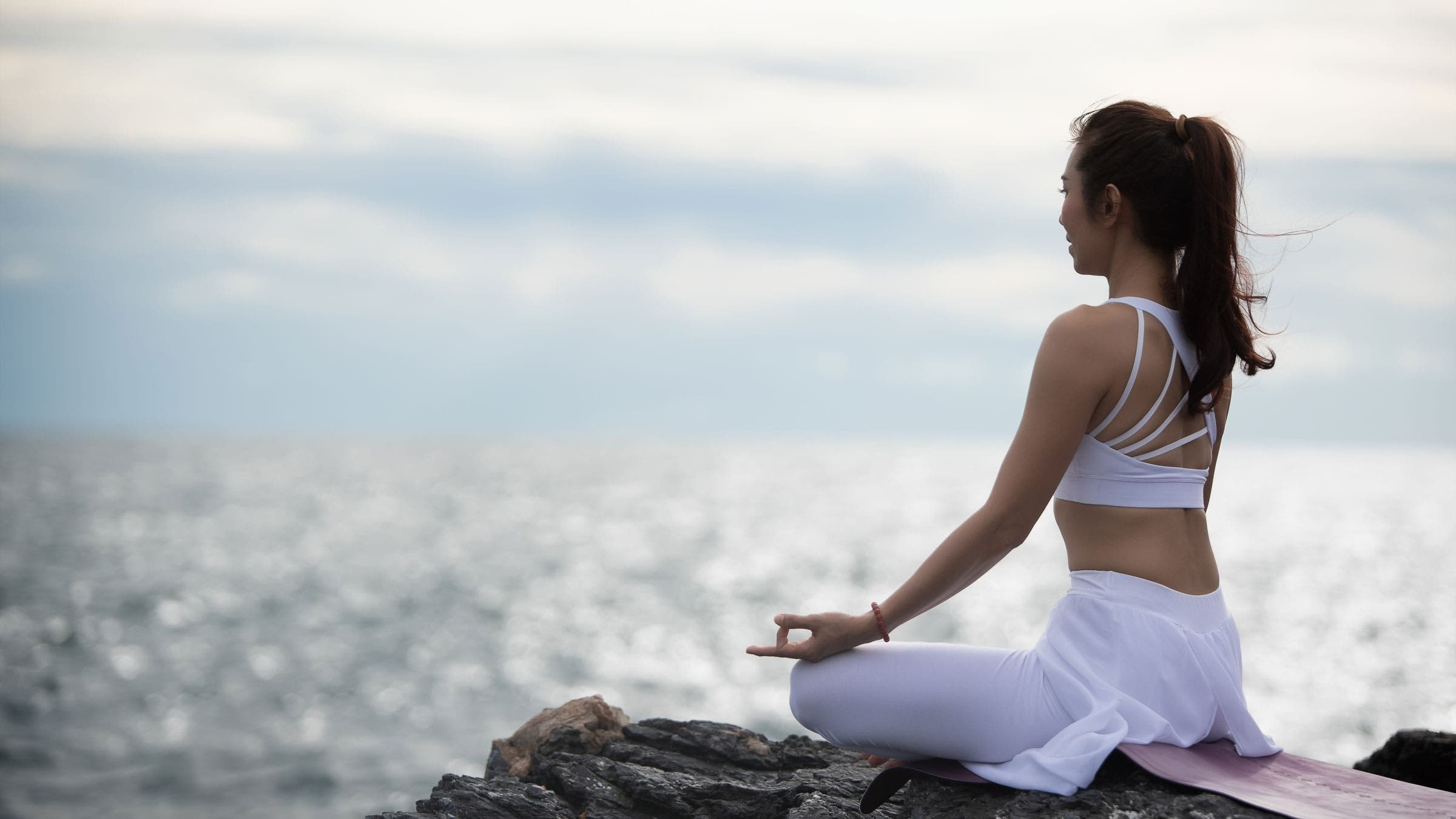 A woman sits and practices a mantra meditation on a rock overlooking the ocean