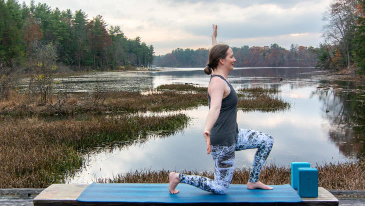 Anna Engels in kneeling lunge with arms wide at a diagonal. Stream and woods in the background