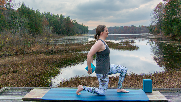 Anna Engels in low lunge with a block behind her hips, against a backdrop of stream and trees