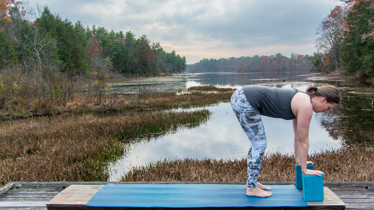 Anna Engels in forward fold with hands on blocks against a backdrop of stream and trees