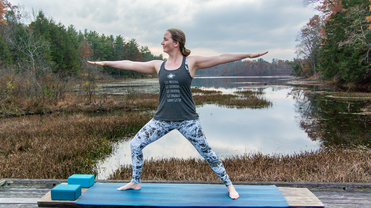 Anna Engels in Warrior II against a backdrop of stream and woods