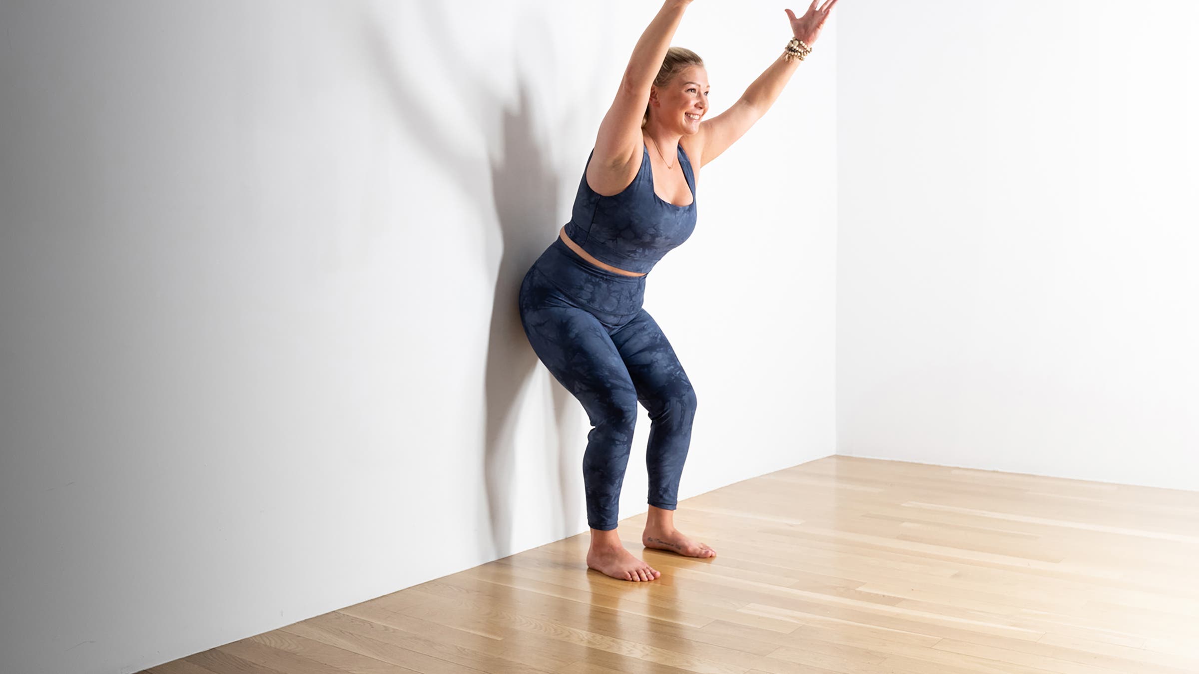 Woman in a chair pose modification against a wall, with arms raised