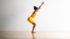 Una mujer con ropa deportiva amarilla realiza una postura de silla de yoga sobre un piso de madera con las rodillas dobladas y los brazos extendidos hacia el techo.