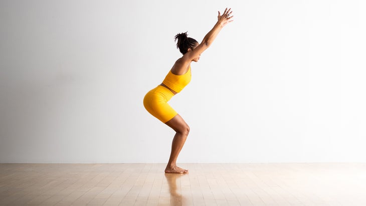 Woman in yellow workout outfit on hardwood floor performing Chair Pose in yoga with her knees bent and arms reaching up toward the ceiling.