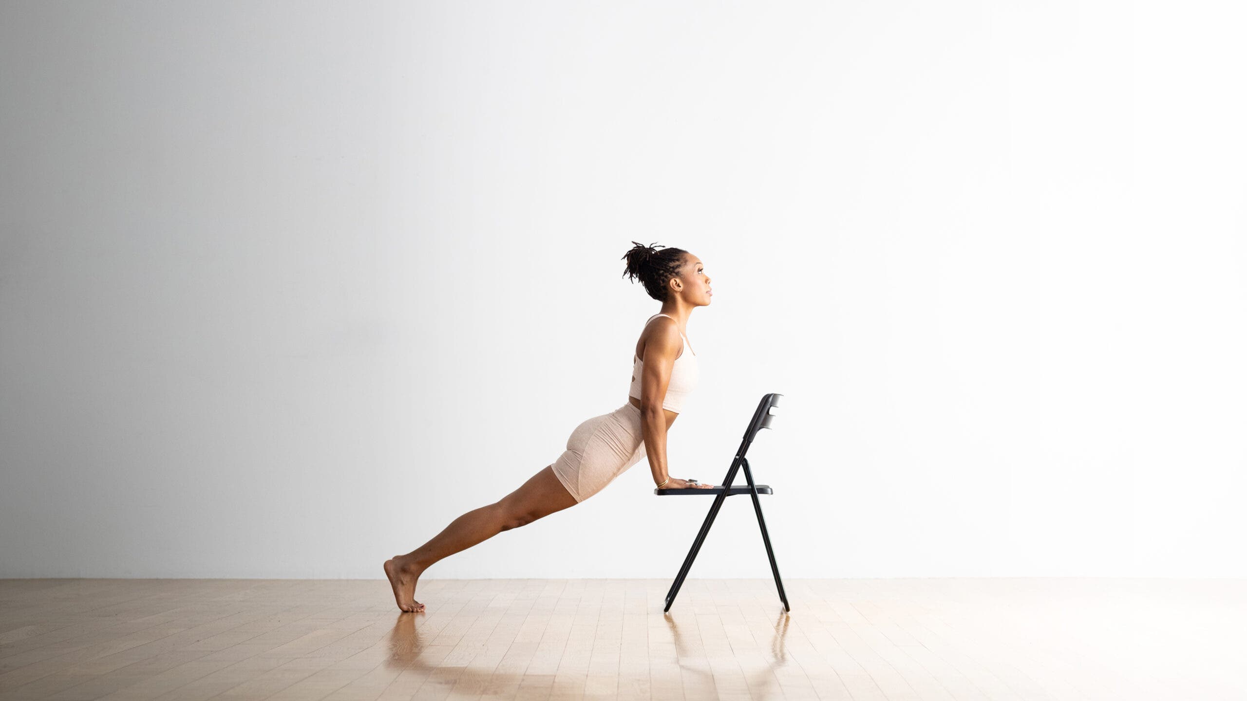 A woman practices Upward Facing Dog with her hands on the seat of a folding chair. She is a Black woman wearing off-white yoga shorts and a matching top. The room has a wood floor and the wall behind her is white.