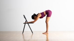 Woman doing a forward bend with her forearms on a black folding chair.