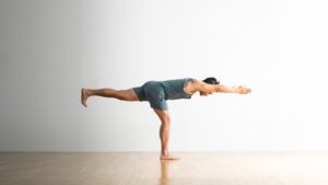 Man in blue workout outfit performing a Warrior III pose on hardwood floor.