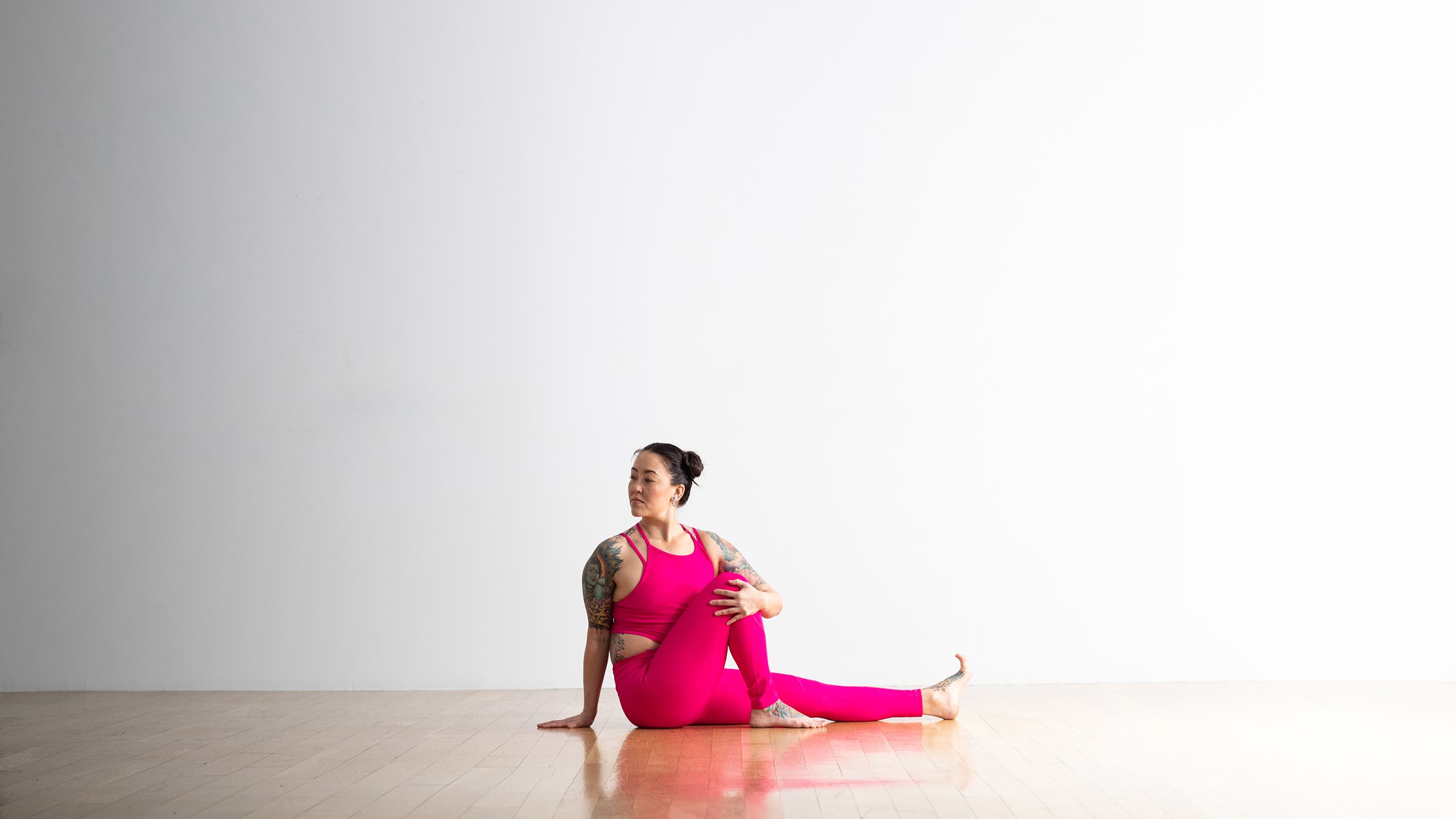 Asian American woman in bright pink top and tights practiced Marichyasana Pose. She is sitting on a hardwood floor with her left leg extended and her right knee bent. She is twisting her torso toward her right leg