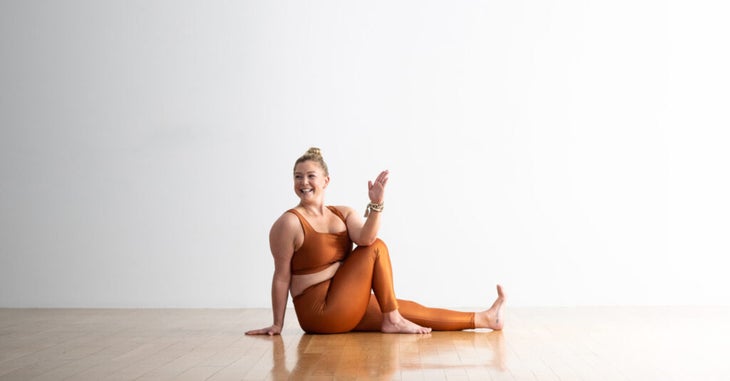 Woman practices Marichyasana III (Marichy's Twist) with her elbow on the outside of her bent knee. She has blonde hair and wears copper-colored yoga tights and matching top. She is in a room with a wood floor and white walls