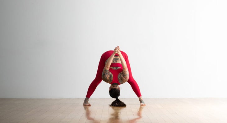 Soozie Kinstler practices a variation of a Wide-Legged Standing Forward Fold. She clasps her hands behind her back and lifts them away from her body and toward the ceiling.