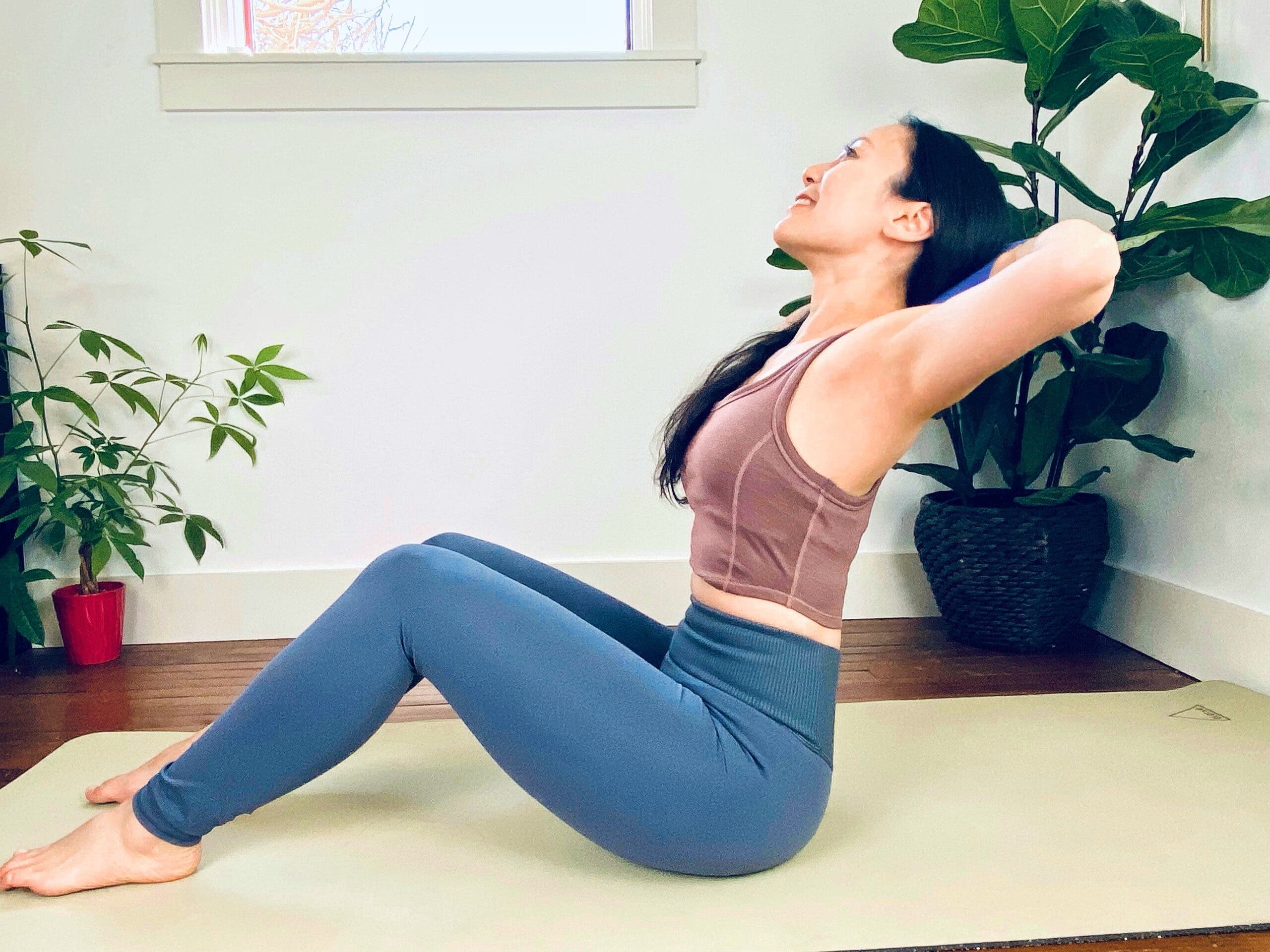 An Asian woman practices Navasana Pose with a yoga block behind her head. She wears a brown top and blue tights, and is sitting on a light green yoga mat. She is in a white room with large plants in the background.