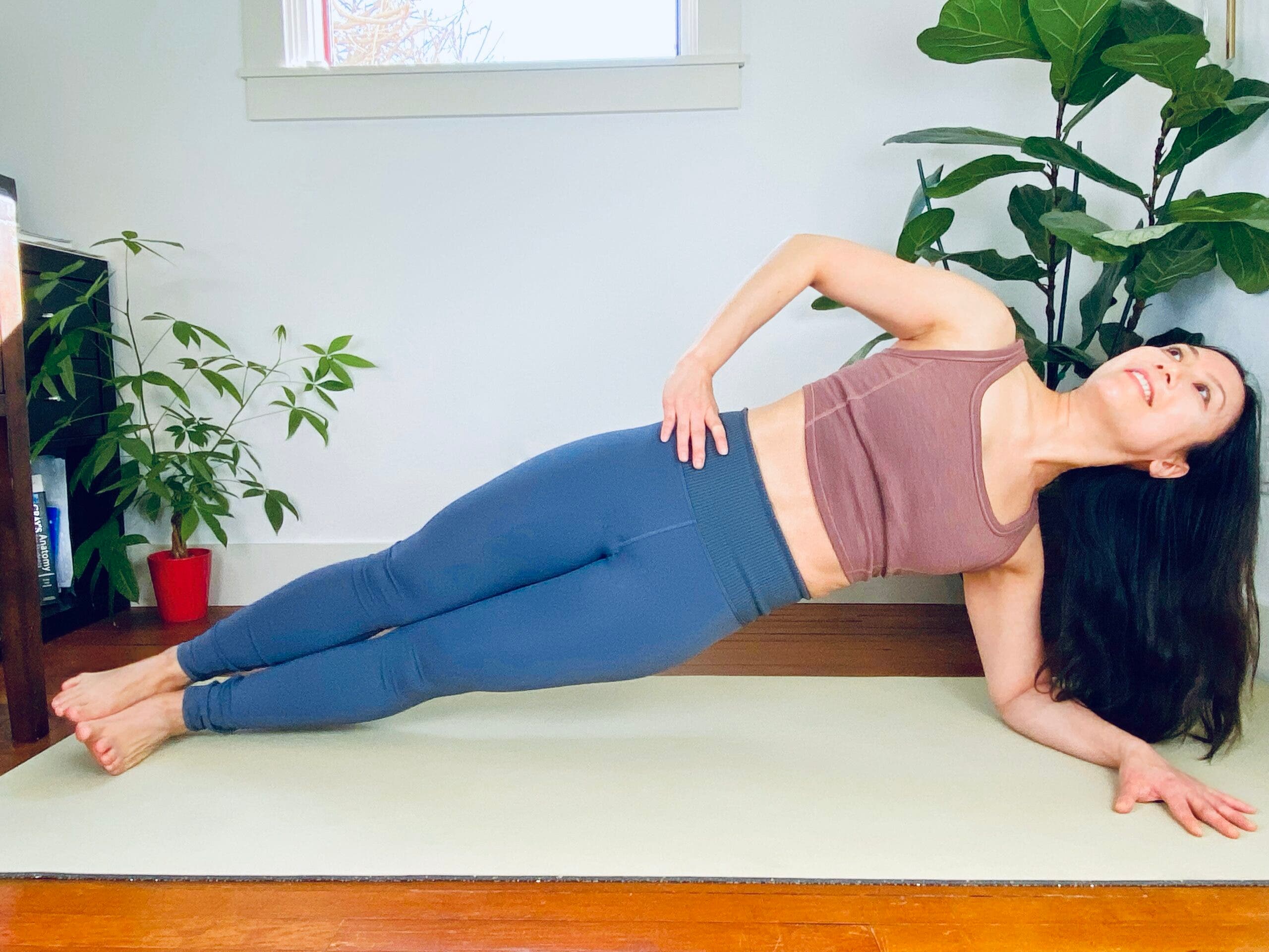 An Asian woman practices side plank pose. She wears a brown top and blue tights, and is on a light green yoga mat. She is in a white room with large plants in the background.