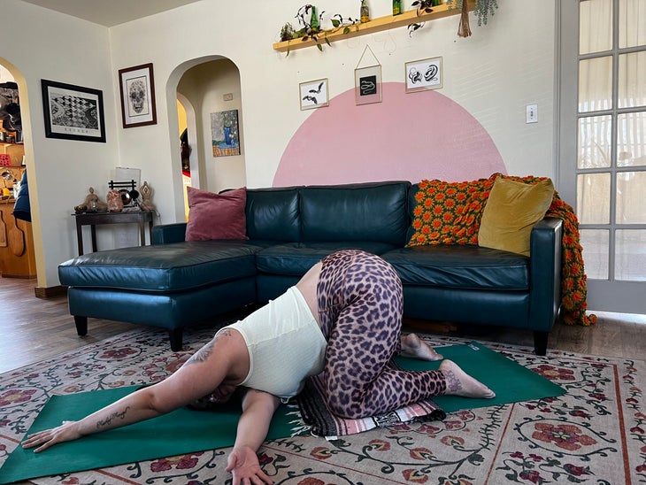 Reyna Cohan kneels to do thread the needle pose. She is wearing leopard-print leggings and a light green top that shows her tattoos. She is sitting on a green yoga mat over a patterned rug. Behind her is a black leather sofa with a yellow-gold velvet pillow and a green and orange afghan.