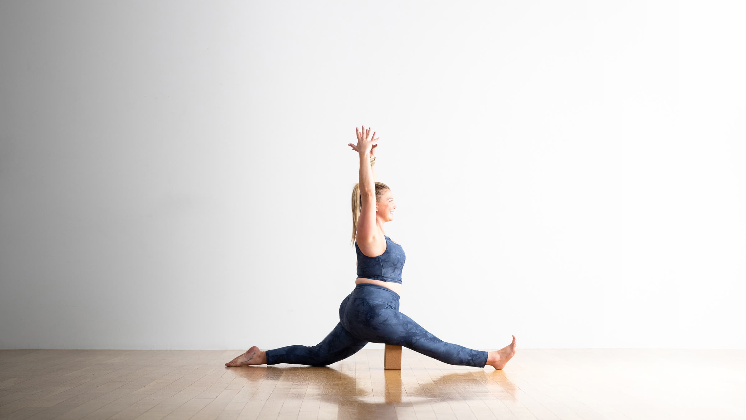 A woman practices Hanumanasana (Monkey Pose) with a block under her front thigh. She is blonde, wearing blue yoga tights and a cropped top of the same colore 