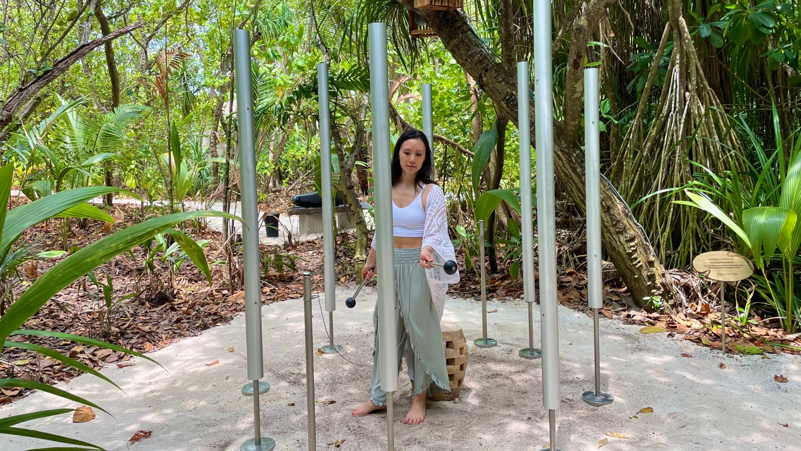 Ingrid Yang plays chimes on the nature walk on the Joali Being Resort in the Maldives. She is surrounded by large metal chimes planted in the white sand. In the background is tropical foliage.