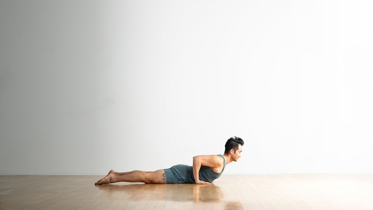 Man with dark hair practices Cobra Pose on a wood floor. The background is white. He is wearing light blue clothes.