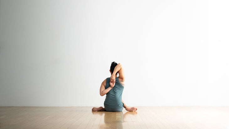 Person sits with his back facing the viewer, right arm reaching down behind shoulder blades and left arm reaching up the back. His hands are clasped together. He is sitting cross legged on a wood floor with a white background.