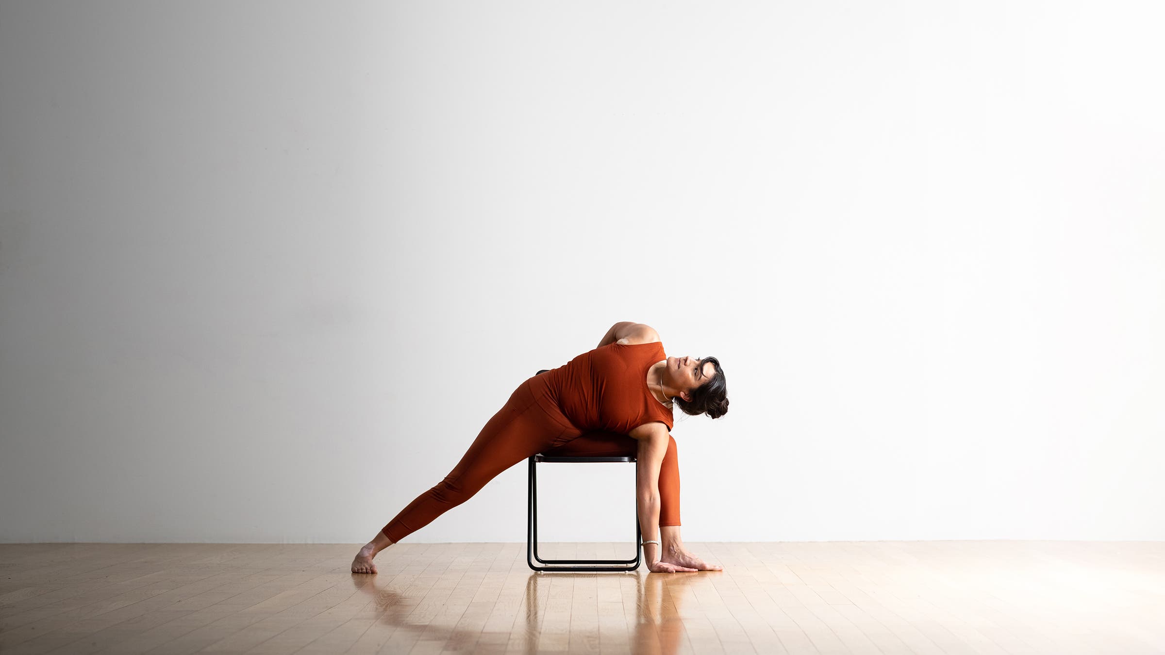 Dark-haired woman in rust colored tights and top practices Extended Side Angle in a chair