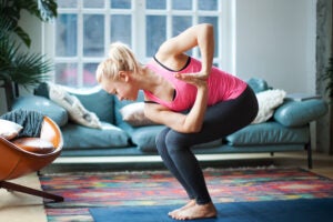 Woman bending her knees and twisting her arms with a green couch in the background.