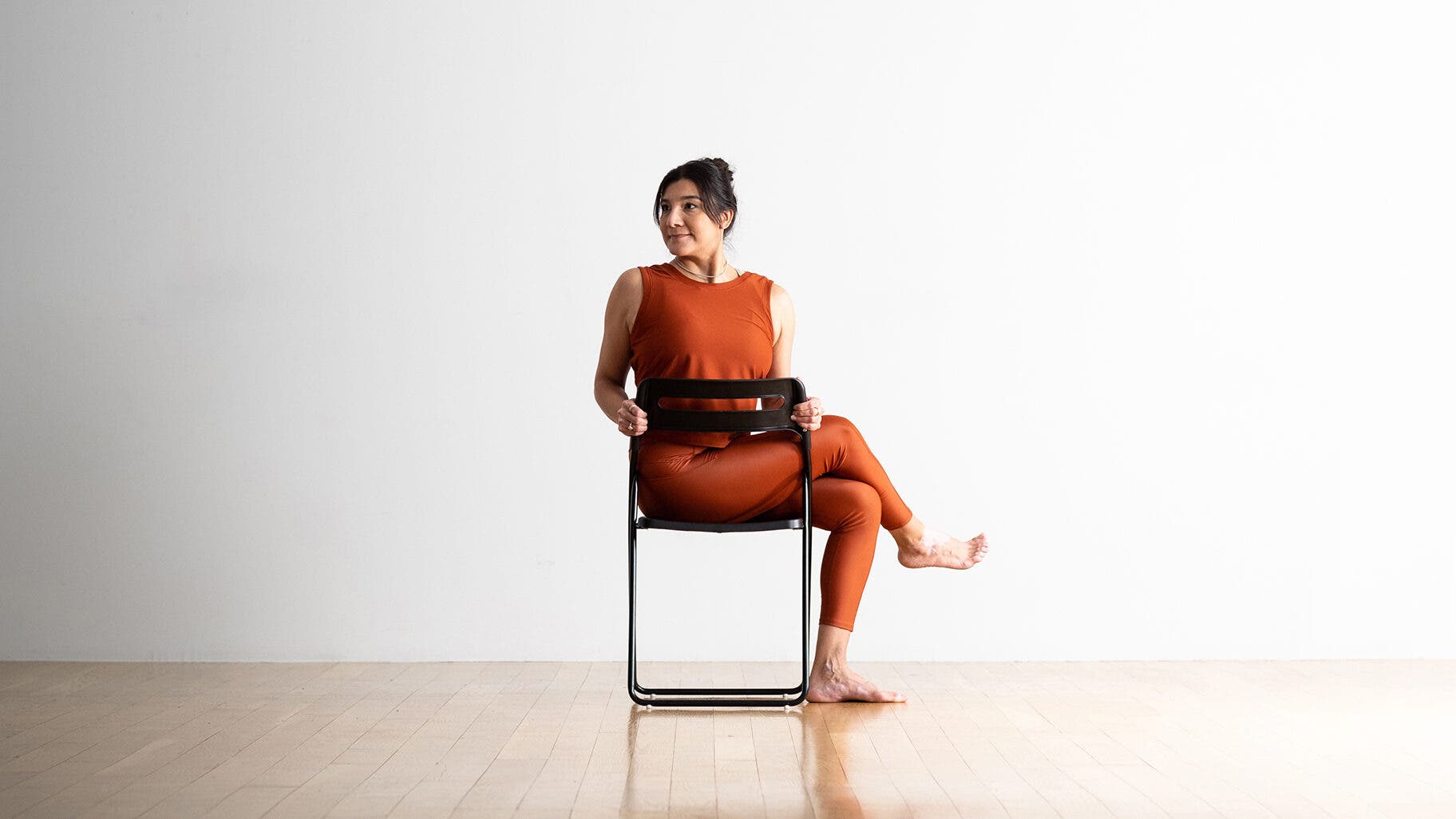 Dark haired woman practices chair yoga by sitting in a version of Half Lord of the Fishes with her chest turned to one side and her legs crossed.