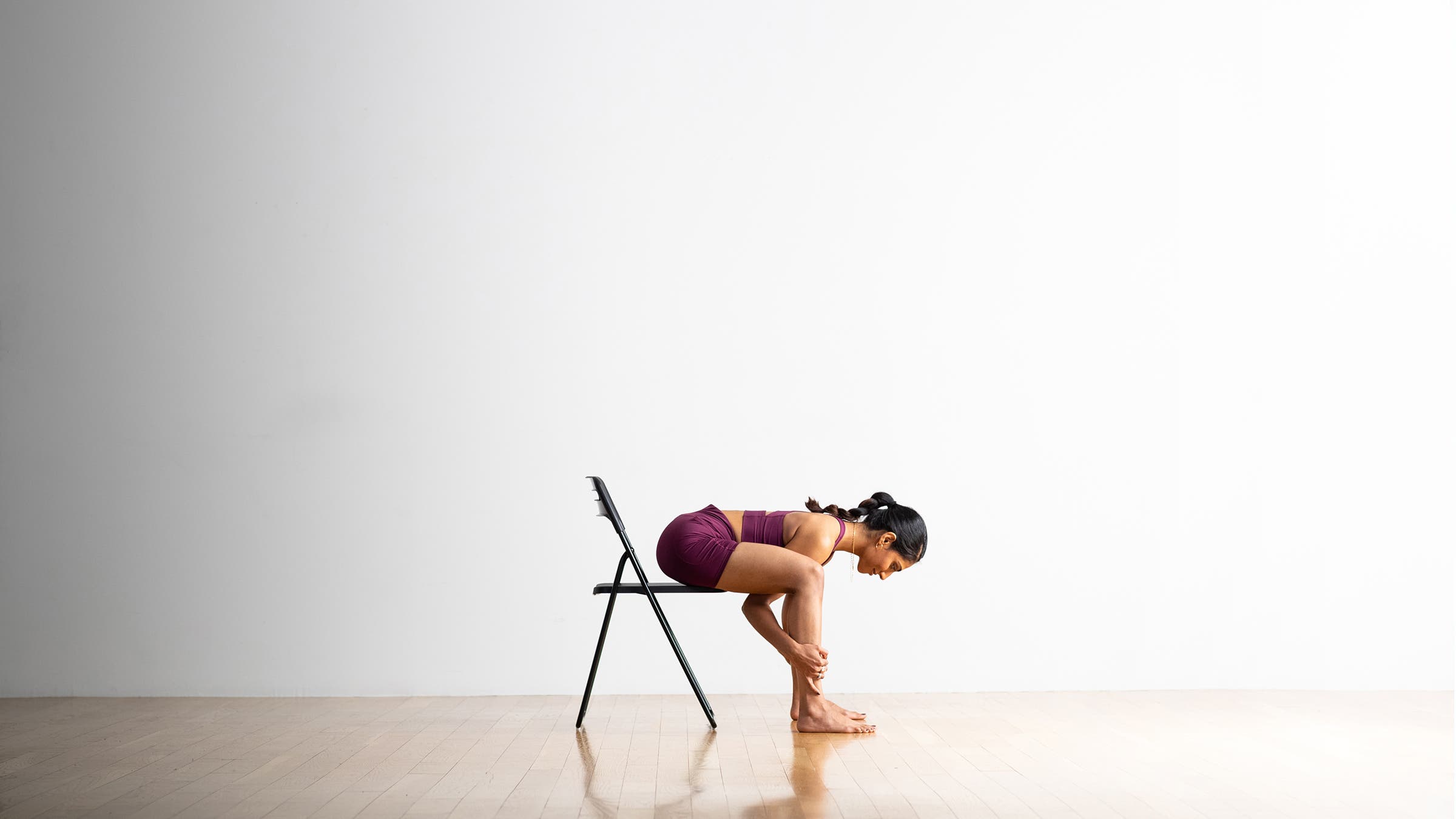 South Asian women bends from the waist to do a modification of Happy Baby pose. She is wearing purple-ish yoga shorts and a matching bra top. The floor is wood and the wall behind her is white.