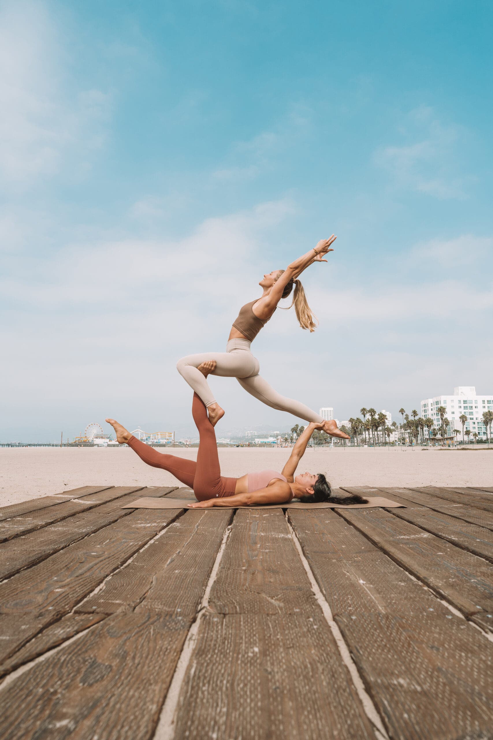 Two women demonstrate Acro Yoga on a beach boarwalk. A woman with dark hair lies on a tan mat. With one leg an one hand, she lifts and holds up another woman in a Crescent lunge position.