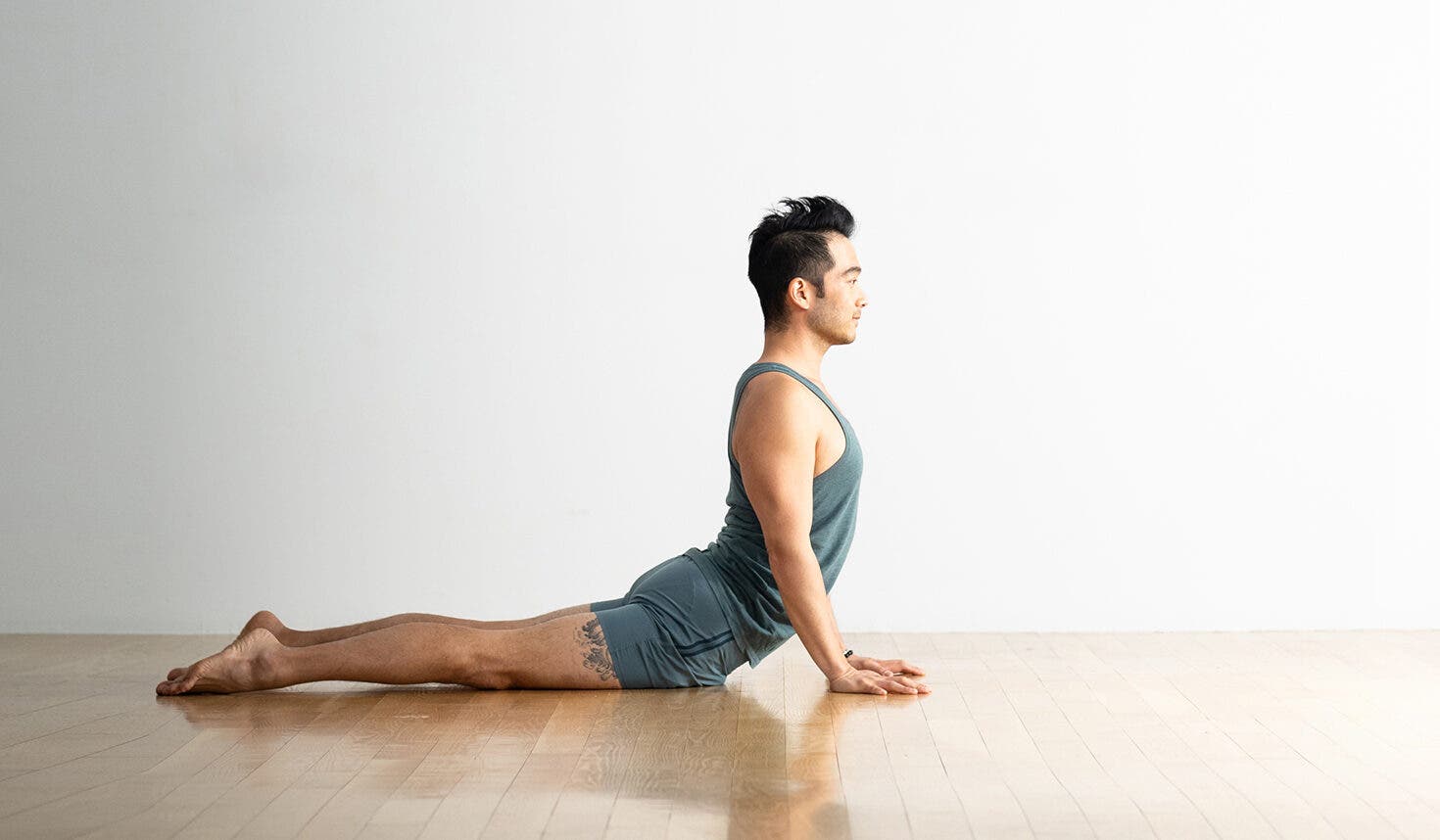Man with dark hair practices Cobra Pose on a wood floor. The background is white. He is wearing light blue clothes.