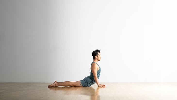 Man with dark hair practices Cobra Pose on a wood floor. The background is white. He is wearing light blue clothes.