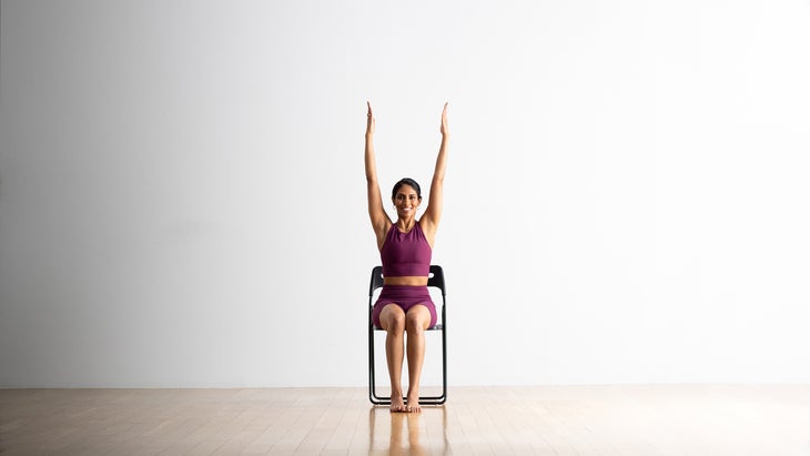 South Asian women sitting in a black chair extending her arms up in a chair yoga pose version of Urdhva Hastasana or Upward Salute