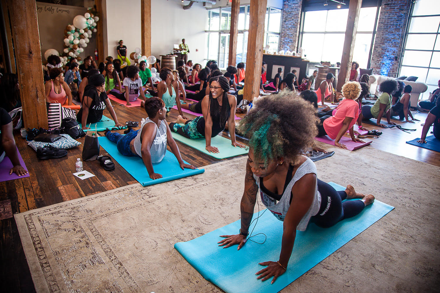 BRITTENY FLOYD-MAYO leads a trap yoga class. She demonstrates a Cobra variation on a light blue mat. Behind her, a large group of mostly African-American women laugh as they practice the pose. They are in a large room with big windows and dark wood floors. 