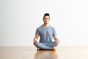 Hiro Landazuri, a man with dark hair, sits cross legged in Easy Pose (Sukhasana) on a light wood floor against a white wall. He is wearing a light blue T-shirt and pants.