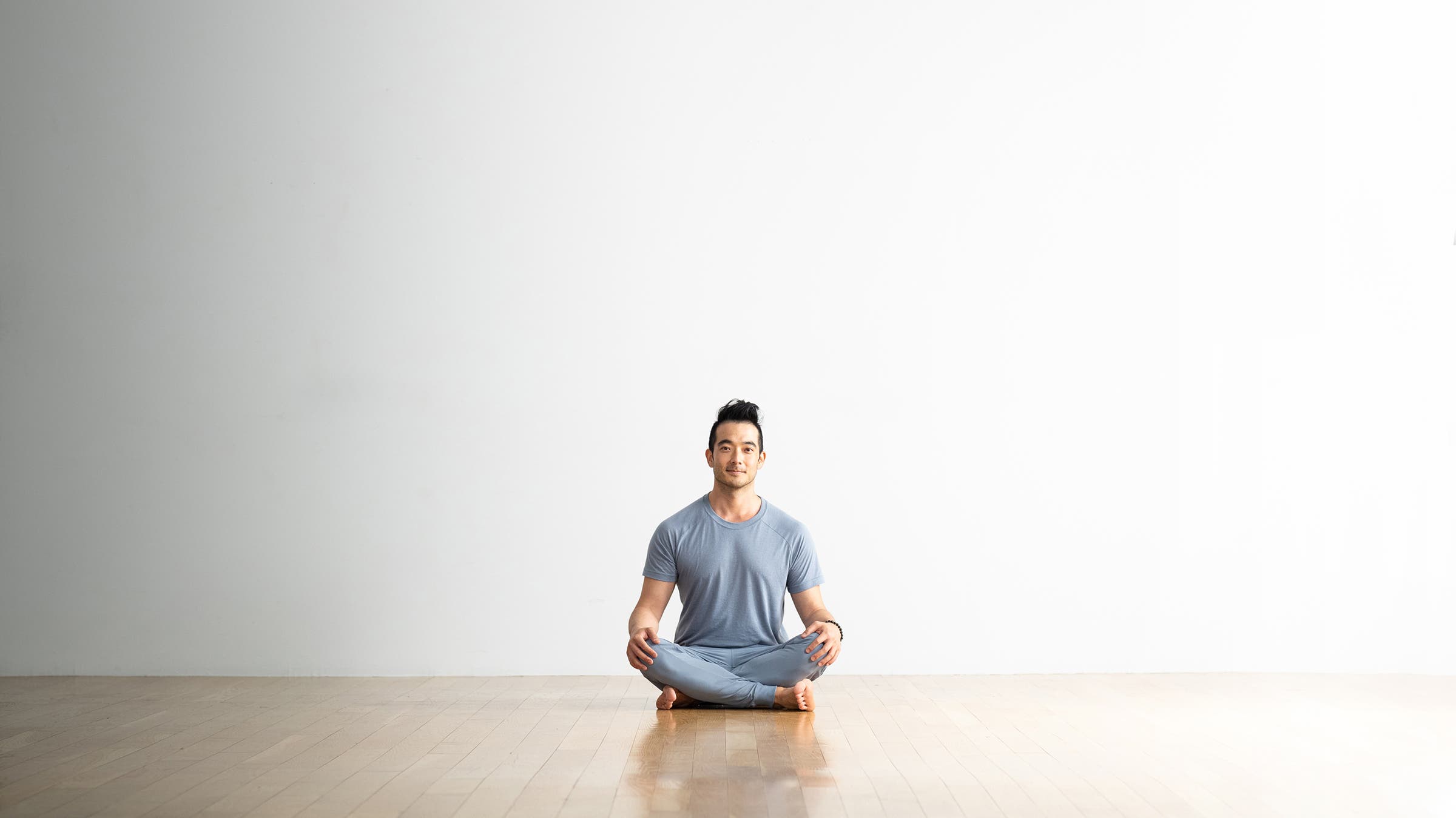Hiro Landazuri, a man with dark hair, sits cross legged in Easy Pose (Sukhasana) on a light wood floor against a white wall. He is wearing a light blue T-shirt and pants.