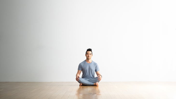 Hiro Landazuri, a man with dark hair, sits cross legged in Easy Pose (Sukhasana) on a light wood floor against a white wall. He is wearing a light blue T-shirt and pants.