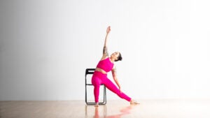 A woman practices Gate Pose sitting in a chair. She has dark hair in two buns, and she is wearing bright magenta yoga pants and cropped top