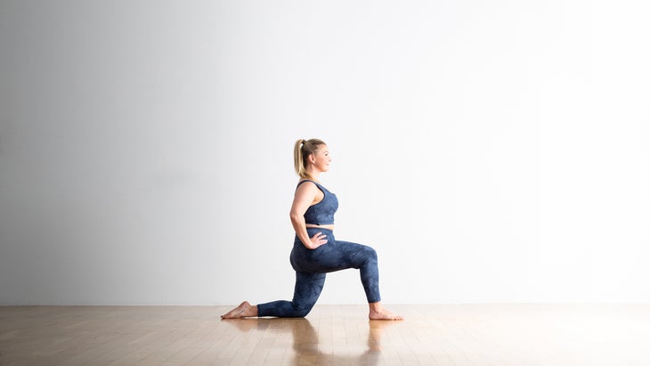 A woman with blond hair kneels in a Low Lunge variation. Her right leg is forward with her foot planted on the floor while kneeling on her left knee. Her hands are on her hips. She is smiling an wearing a blue pattern crop top and matching pants. The wall behind her is white, the floor looks like light hardwood