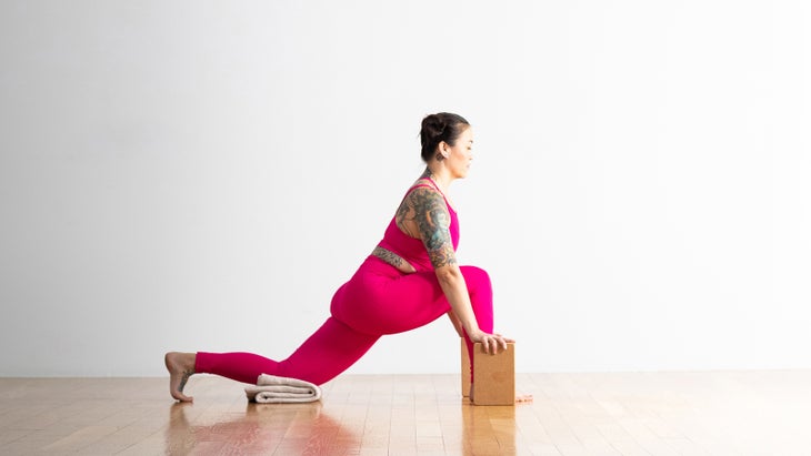 A woman practices a lunge pose. Her right foot in forward; her left knee is resting on a folded blanket. She has her hands on cork bloks. She has on red leggings and a cropped top.