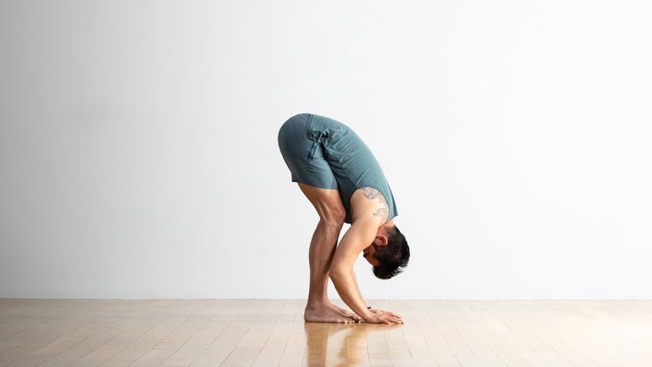 A man with dark hair bends forward in Uttanasana, Standing forward fold. He wears gray-blue shorts and top. His knees are slightly bent. He has his hands on the hardwood floor near his feet.