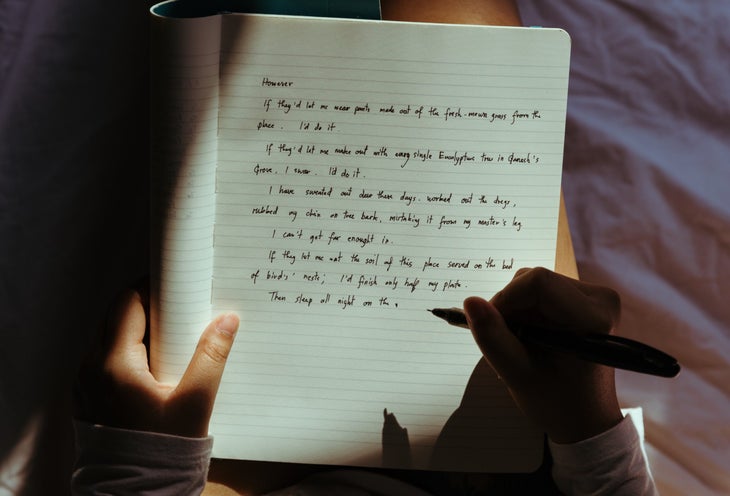 A person holds a notebook with their pen poised above the page. Light shines down on the black writing.