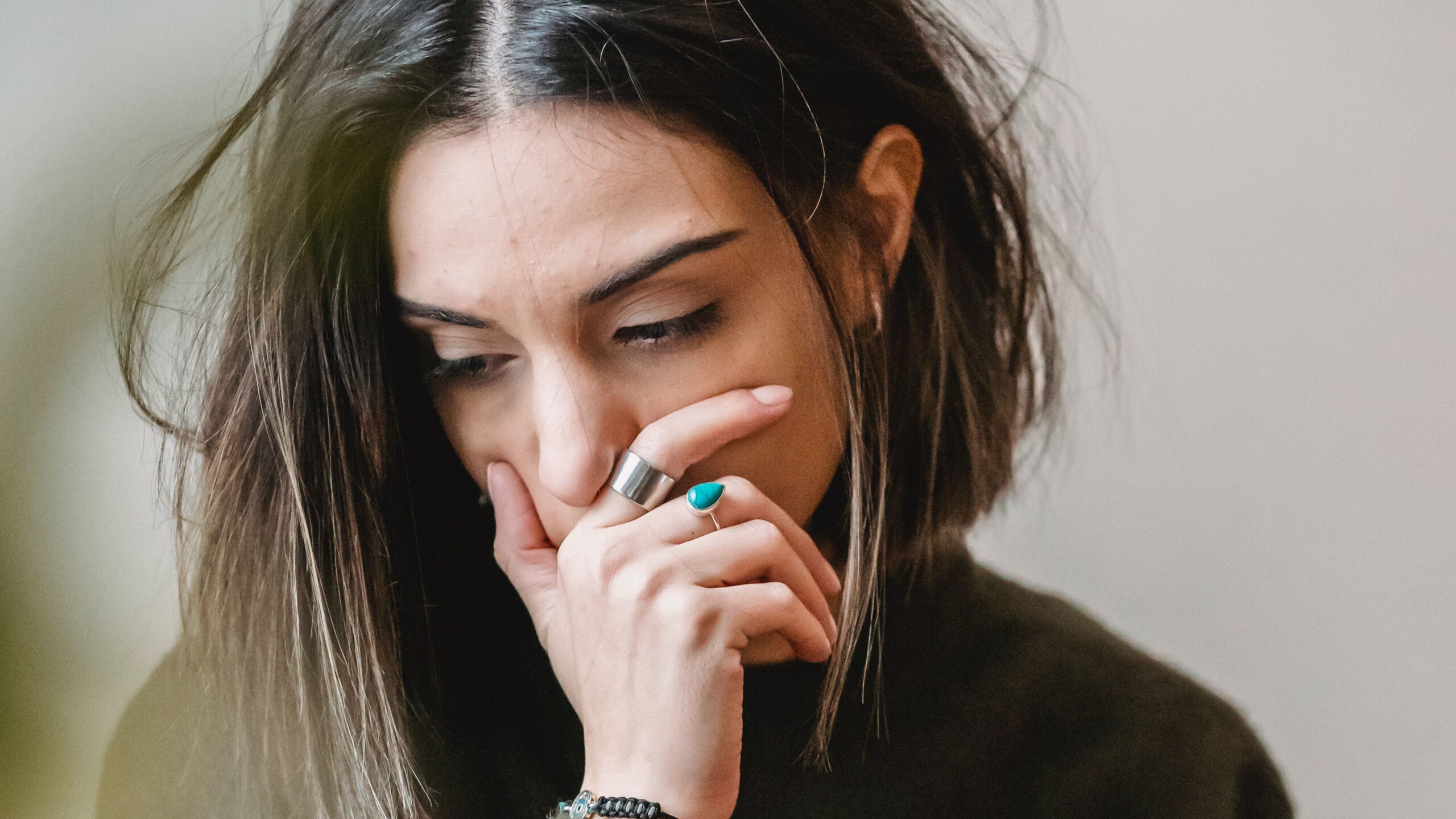 A person with straight, brown hair cropped in a bob holds her hand over her mouth. She looks sad, ill, or worried. She wears a metal ring on her first finger and a blue ring on her middle finger.