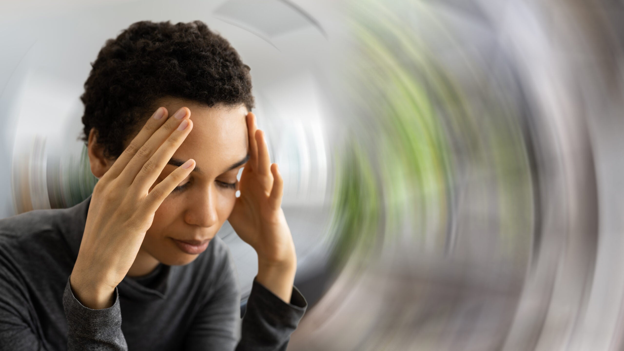 A black woman with short curly hair has her hands to her forehead. The background around her is blurred and swirling or spinning.