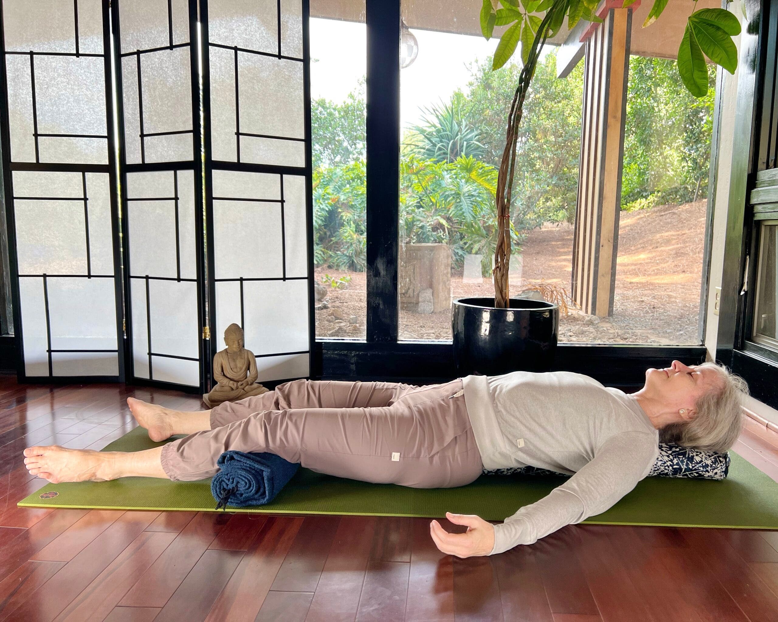 A woman with white hair practices Savasana, Corpse Pose. She is lying on a green mat on a brown wood floor, wearing brown pants and a light brown shirt. In the background are floor-to-ceiling windows with a view of trees. A shoji screen and a small Buddha statue are on the left and a tall plant is behind her. She is supported by a bolster under her head.