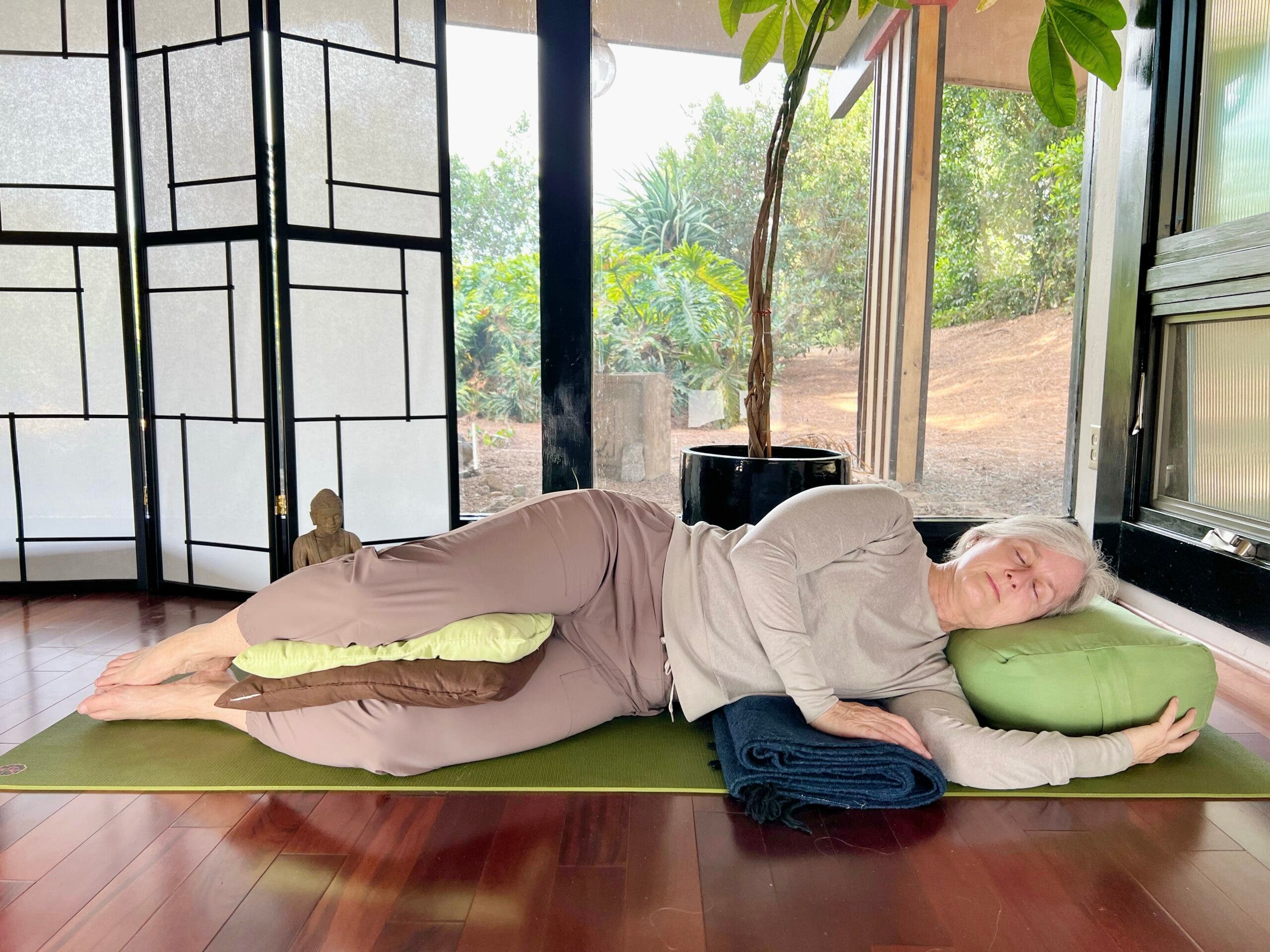 A woman with white hair practices Savasana, Corpse Pose. She is lying on a green mat on a brown wood floor, wearing brown pants and a light brown shirt. In the background are floor-to-ceiling windows with a view of trees. A shoji screen and a small Buddha statue are on the left and a tall plant is behind her. She is lying on her side with pillows under her head and between her knees.