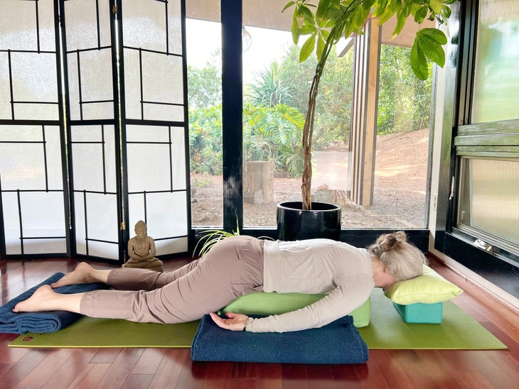 A woman with white hair practices Savasana, Corpse Pose. She is lying on a green mat on a brown wood floor, wearing brown pants and a light brown shirt. In the background are floor-to-ceiling windows with a view of trees. A shoji screen and a small Buddha statue are on the left and a tall plant is behind her. She is lying face down with pillows and blankets under her body and her head resting on a block and pillow.