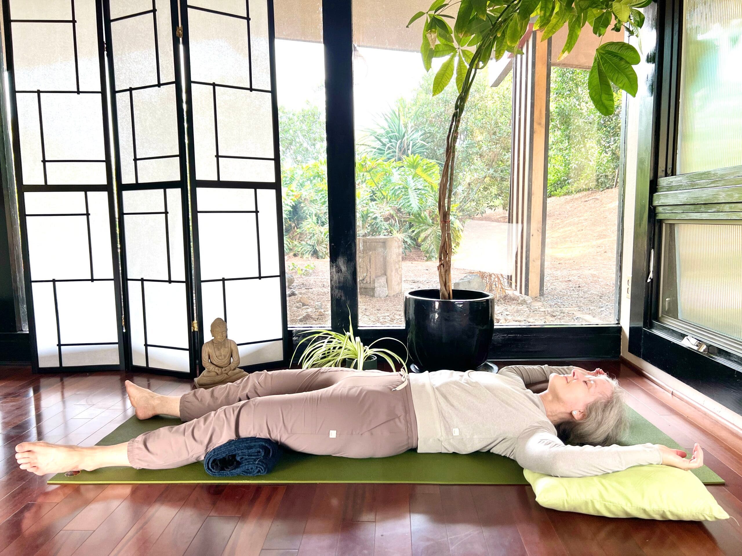 A woman with white hair practices Savasana, Corpse Pose. She is lying on a green mat on a brown wood floor, wearing brown pants and a light brown shirt. In the background are floor-to-ceiling windows with a view of trees. A shoji screen and a small Buddha statue are on the left and a tall plant is behind her. She is lying on her back with her arms reaching out and upward with green pillows under her arms.