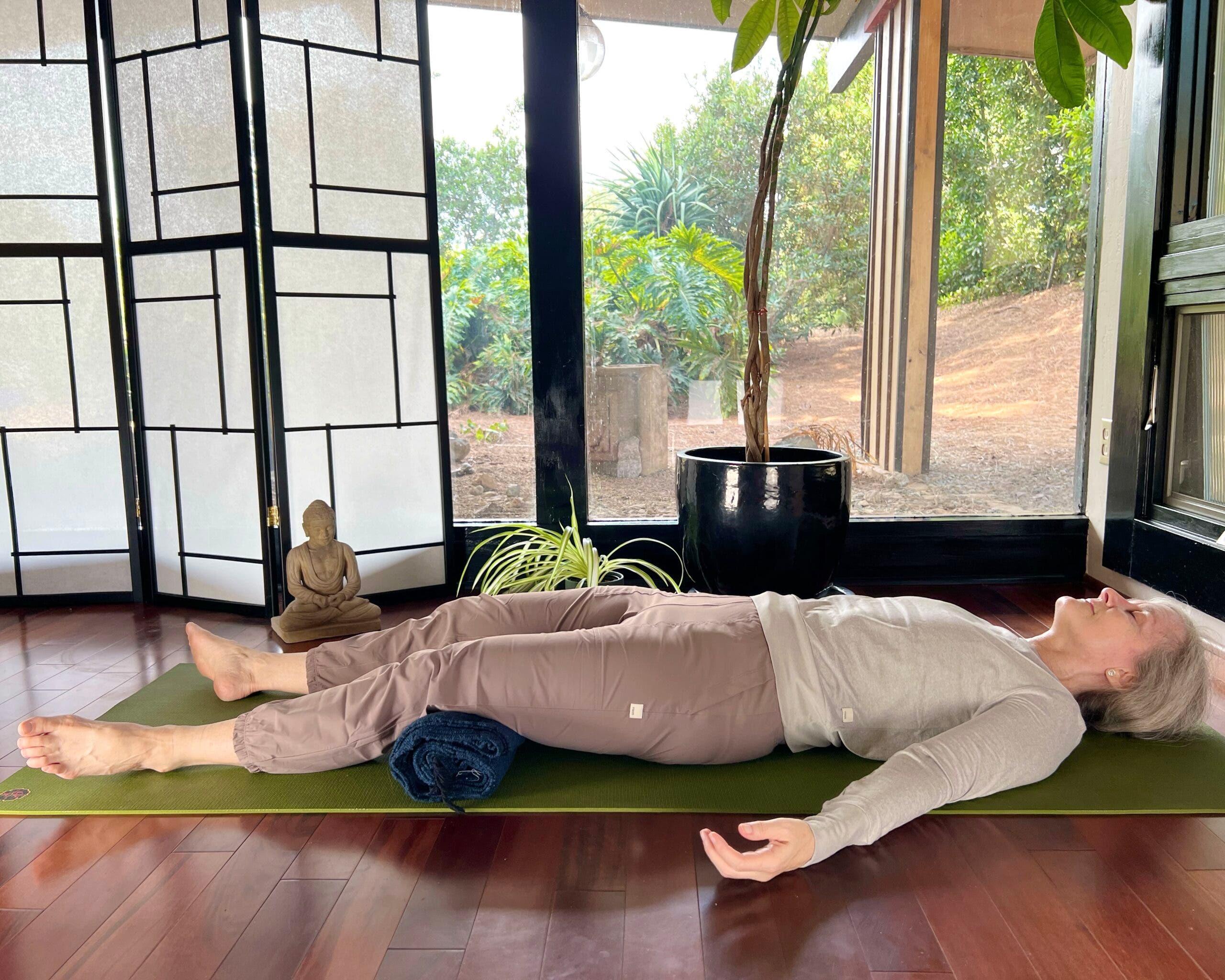 A woman with white hair practices Savasana, Corpse Pose. She is lying on a green mat on a brown wood floor, wearing brown pants and a light brown shirt. In the background are floor-to-ceiling windows with a view of trees. A shoji screen and a small Buddha statue are on the left and a tall plant is behind her.
