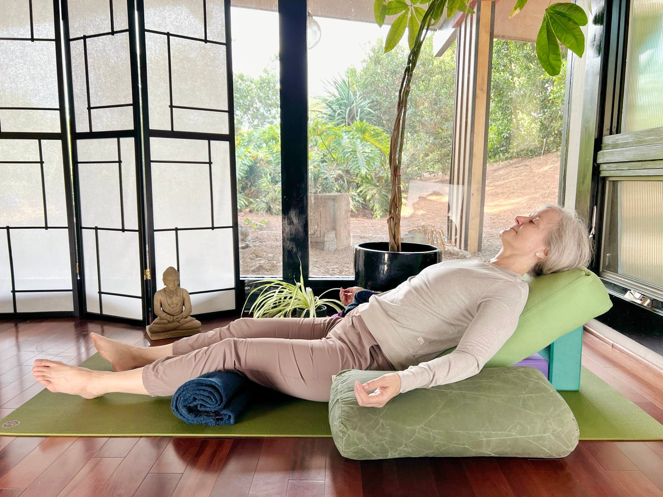 A woman with white hair practices Savasana, Corpse Pose. She is lying on a green mat on a brown wood floor, wearing brown pants and a light brown shirt. In the background are floor-to-ceiling windows with a view of trees. A shoji screen and a small Buddha statue are on the left and a tall plant is behind her. She is lying back on a bolster elevated on blocks. Her arms are on green bolsters.
